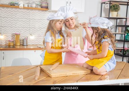 Lächelnde kaukasische Mutter lehrt Töchter, Teig mit Nudelholz in der Küche auszurollen. Hausmannskost-Konzept Stockfoto