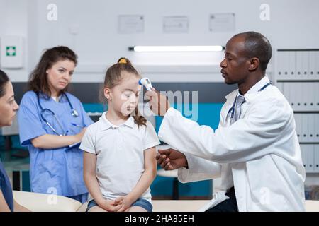 afroamerikanischer Kinderarzt, der ein medizinisches Infrarot-Thermometer auf die Stirn eines Kindes legt, um die Temperatur während der Krankheitsuntersuchung im Krankenhausbüro zu messen. Gesundheitsdienste Stockfoto