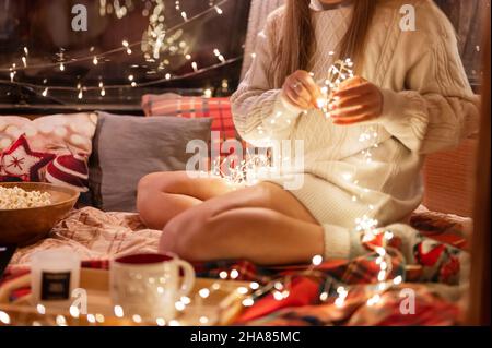Frau in einem gemütlichen bequemen gestrickten Wollpullover hält in den Händen glühende Lichter Girlande und Dekoration Bett in Reise-Anhänger Mobilheim. Neujahr und kri Stockfoto