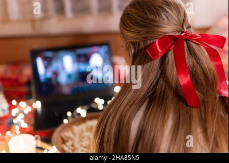 Hinter dem weiblichen Kopf mit langen blondie Haaren und roten Bogen Rückansicht beobachten den weihnachtsfilm auf Laptop auf Camping Home in gemütlichen Bett mit glühenden Lichtern Stockfoto