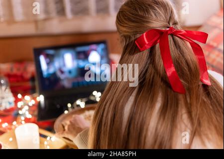 Hinter dem weiblichen Kopf mit langen blondie Haaren und roten Bogen Rückansicht beobachten den weihnachtsfilm auf Laptop auf Camping Home in gemütlichen Bett mit glühenden Lichtern Stockfoto