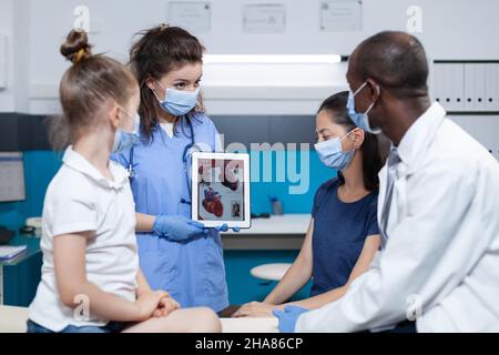 Medizinische Versorgung mit einer Tablette mit Herzkompetenz während der klinischen Verabredung im Krankenhausbüro auf dem Bildschirm. afroamerikanischer Kinderarzt mit schützender Gesichtsmaische gegen Coronavirus Stockfoto