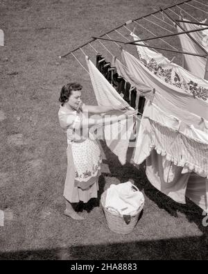 1950S FRAU HÄNGEND WÄSCHE AUF OUTDOOR WÄSCHELEINE IM HINTERHOF TROCKNEN - H2864 DEB001 HARS LÄNDLICHEN HAUS LEBEN KOPIE RAUM LANGE DAMEN TROCKENE PERSONEN PFLEGE B&W HAUSFRAU FRISCHE TRÄUME HAUSFRAUEN WELLNESS FRÖHLICH HIGH-ANGLE CHORE TROCKNEN WÄSCHELEINE HYGIENE HAUSFRAUEN LÄCHELT AUFGABEN FRÖHLICHE DEB001 BETTWÄSCHE MID-ADULT MID-ADULT FRAU AUFGABE SCHWARZ-WEISS KAUKASISCHEN ETHNIE ALTMODISCH Stockfoto