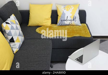 Wohnzimmer verfügt über ein silbernes Macbook Pro, das auf dem Tisch durch ein bequemes graues Sofa mit gelben und gemusterten Kissen geöffnet wird Stockfoto