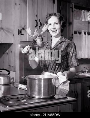 1950S 1960S LÄCHELNDE FRAU HAUSFRAU, DIE BEIM KOCHEN DER KAMERA BEIM ABENDESSEN DABEI IST, HUMMER IN EINEN TOPF MIT KOCHENDEM WASSER ZU GEBEN HERD - H3314 DEB001 HARS HOME LIFE LUXURY VORBEREITUNG HALBLANGE DAMEN PERSONEN ÜBER AUSDRÜCKE B&W BLICKKONTAKT HAUSFRAU SETZEN HAUSFRAUEN FRÖHLICHE WAHL IN VON AUF ZU HAUSFRAUEN LÄCHELT KONZEPTUELL KOCHEND FRÖHLICH STILVOLLE DEB001 JUNGE ERWACHSENE FRAU SCHWARZ UND WEISS KAUKASISCHE ETHNIE ALTMODISCH Stockfoto