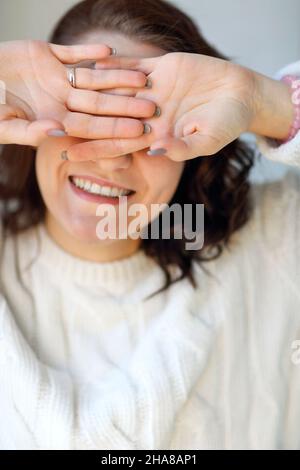 Fröhliche, entzückende Frau in Strickpullover, die ihre Augen mit den Handflächen nach außen schließt und vor der Kamera lächelt, während sie Freizeit zu Hause verbringt, fröhliches FEM Stockfoto