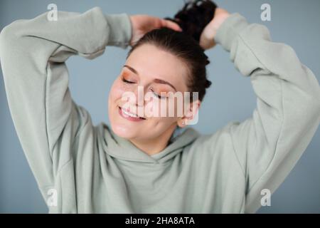 Zufriedene entzückte Frau mit Glitzer Make-up, trägt lässiges gemütliches Sweatshirt, das ihre Augen mit verträumtem Gesichtsausdruck schließt, glättet die Haare in Ponyta Stockfoto