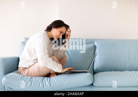 Junge, nachdenkliche Frau in legerer Kleidung sitzt mit ihren Beinen auf dem Sofa und benutzt die Tastatur auf dem Laptop, starrt auf den Computerbildschirm und sucht nach wichtigen notwendigen Stockfoto