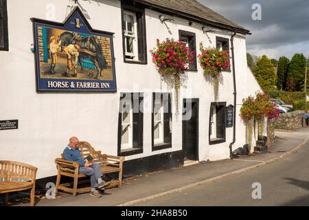 Großbritannien, Cumbria, Allerdale, Keswick, Threlkeld, Mann liest in Sonnenschein unter Horse and Farrier Pub Schild Stockfoto