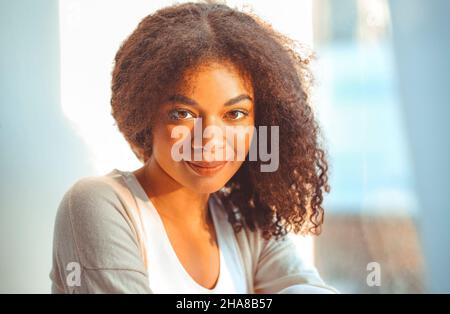 Erfreut entspannte afroamerikanische Ethnie Mädchen in Homewear sitzen auf Fensterbank vor dem Hintergrund der Vorhänge flattern vor leichtem Wind, glücklich afro Stockfoto