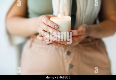 Seitenportrait einer jungen, ruhigen Frau, die eine brennende Kerze in den Händen hält, die Augen schließt und ihr Gesicht näher bringt und ihr Aroma einatmet, wobei sie den vertrauten Duft riecht Stockfoto