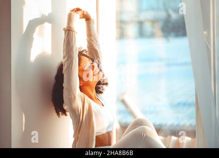 Erfreut entspannte afroamerikanische Ethnie Mädchen in Homewear sitzen auf Fensterbank vor dem Hintergrund der Vorhänge flattern vor leichtem Wind, glücklich afro Stockfoto