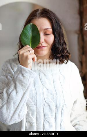 Portrait eines jungen Brünetten Mädchens mit schön gestylten Haaren in weißem warmen Strickpullover, ruhig steht und bedeckt ihr ein Auge mit grünem Pflanzenblatt, Stockfoto