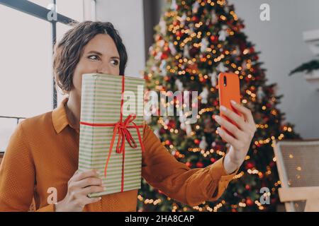 Fröhliche spanische helle Brünette Dame macht Selfie auf dem Smartphone, während posiert mit Weihnachten Geschenk Stockfoto