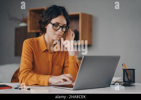 Eine versetzte Spanierin in einer Brille sitzt am Schreibtisch und tippt während der Fernarbeit zu Hause auf dem Laptop Stockfoto