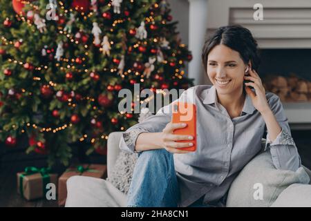 Glückliche hispanische Frau in schnurlosen Ohrhörern, die das Telefon in der Nähe des Weihnachtsbaums zu Hause hielt Stockfoto