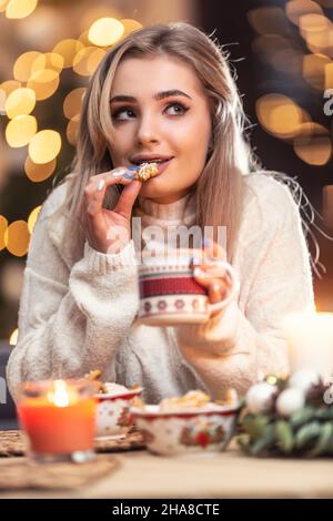 Eine schöne junge blonde Frau schmeckt frischen Weihnachts-Lebkuchen mit großartigem Geschmack und hält eine Tasse Bunch Tee oder Kaffee. Stockfoto