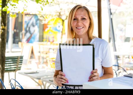 Erwachsene blonde Frau, die lächelt und in einem Restaurant sitzt, während sie wählt, was sie von der Speisekarte bestellt, Platz kopiert und sich vorstellt Stockfoto