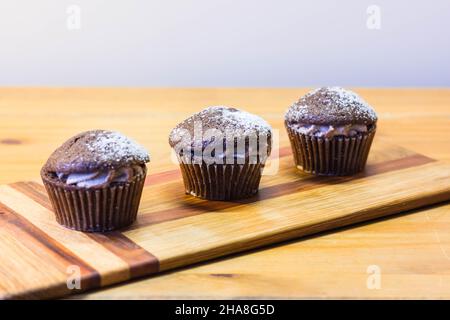 Drei Schokoladen-Victoria-Sponge-Mini-Cup-Kuchen mit Kakaocreme gefüllt und mit Zucker auf Kirschholzbrett bestäubt Stockfoto