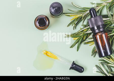 Rosmarin ätherisches Öl fließt aus der Pipette neben Flaschen und frischen Rosmarinkräutern auf grünem Hintergrund, Draufsicht Stockfoto