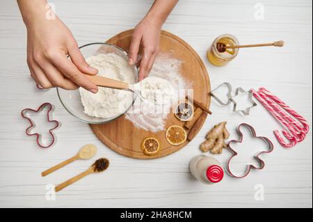 Flache weibliche Hände, die einen Löffel mit Mehl über einer Schüssel auf einem Holztisch mit getrockneten Orangenscheiben und Schneideformen halten Stockfoto