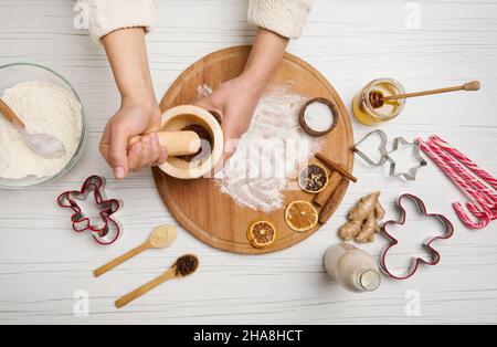 Aus der Perspektive sehen Sie, wie die Hände der Frau Gewürze und Zutaten in einem Marmormörtel schleifen, während Sie Weihnachtskekse auf dem Küchentisch zubereiten Stockfoto