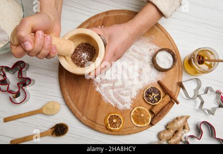 Frauenhänden, die Zutaten in Marmormörtel auf einem Holzbrett mahlen, das mit Mehl und getrockneten Orangenscheiben bestreut wurde, Zimtstangen darauf liegend, geschnitten Stockfoto