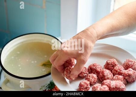 Die Hand einer Frau nimmt rohe Fleischbällchen von einem Teller und wirft sie in einen Topf kochender Suppe. Gesunde Hausmannskost. Stockfoto