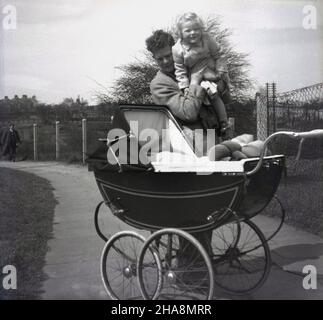 1950s, historisch, draußen auf einem Pfad in einem Park, hält ein Vater seine junge Tochter hoch, neben einem traditionellen Kinderwagen aus der Zeit, in dem sein anderes neugeborenes Kind liegt, in das das Mädchen hineinsehen will, England, Großbritannien. Stockfoto