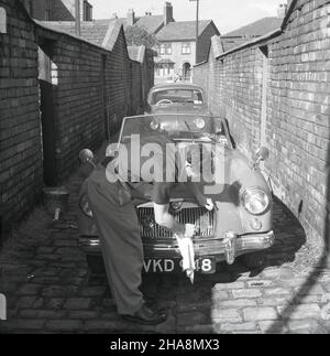 1958, historisch, in einer engen, gepflasterten Backsteingasse hinter einigen viktorianischen Reihenhäusern, putzt ein Mann seinen oben offenen MG-Sportwagen, England, Großbritannien. Neben dem Auto ein Zinneimer mit Wasser. In dieser Zeit war MG (Morris Garagen) eine britische Marke, die Sportwagen produzierte, und der MG MGA war ein Sportwagen aus den Jahren 1955 bis 1962. Das Auto hier, eine MG MGA Twin Cam, wurde von 1958 bis 1960 produziert. Stockfoto