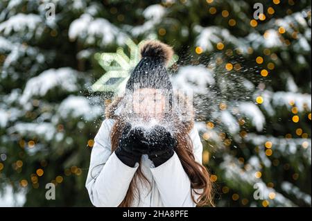 Die junge Frau bläst Schnee aus ihren Händen, während sie vor dem Hintergrund eines Weihnachtsbaums steht Stockfoto