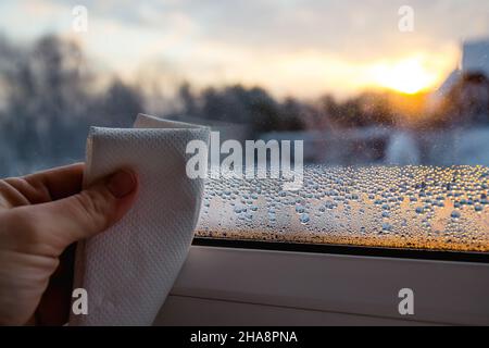 Nahaufnahme der Hand eines Menschen mit einem Papiertuch, das nasse Kondenswassertropfen am kalten Wintermorgen bei Sonnenaufgang aus dem Glasfenster trocknet. Stockfoto