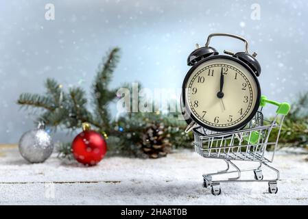 Weihnachtseinkaufskonzept. Wecker in einem Einkaufswagen neben dem Tannenzweig, während es auf einem Holztisch schneit Stockfoto