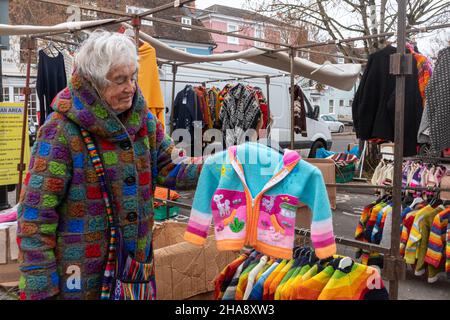 Eine ältere Frau, die an einem Marktstand arbeitet, der handgefertigte Wolljacken, Pullover, Strickjacken, UK in leuchtenden Farben verkauft. Marktanbieter. Stockfoto