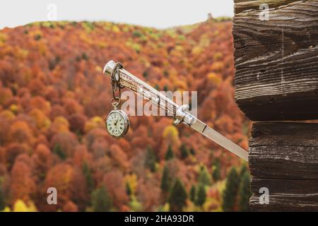 Eine alte Uhr, die an einem Messer hängt, hängt in einem alten Häuschen mit einer herbstlichen Landschaft im Hintergrund Stockfoto