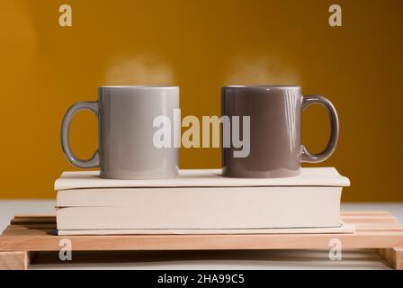 Zwei braune und graue Tassen Kaffee oder Tee mit heißem Dampf auf dem Tisch, mit Buch und braunem Hintergrund Stockfoto