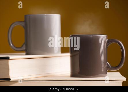 Zwei braune und graue Tassen Kaffee oder Tee mit heißem Dampf auf dem Tisch, mit Buch und braunem Hintergrund Stockfoto