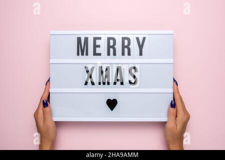 Lightbox mit dem Text FROHE WEIHNACHTEN in den Händen der Frauen auf einem rosa Hintergrund. Minimales Weihnachts- oder Neujahrskonzept. Stockfoto