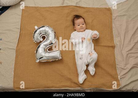 Ein Säugling liegt auf einer Musselin-Decke in der Nähe eines silbernen Folienballons in der Form von 2 und tanzt. Ein glückliches Mädchen in einteiliger Kleidung feiert Stockfoto