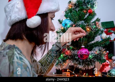 Frau trägt einen Weihnachtsmann Hut, schmücken einen Weihnachtsbaum mit einem vintage rosa festliche Ornament mit einem fröhlichen Weihnachtszeichen. Winterurlaub. Stockfoto