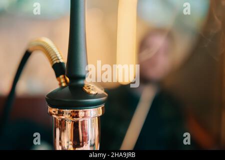 Shisha auf dem Tisch in der Loungebar. Rauchende Person in verschwommenem Hintergrund. Nahaufnahme von schwarzer Pfeife und goldenem Shisha-Kolben. Freizeit im orientalischen Café. Stockfoto