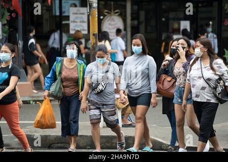 Menschen auf den Philippinen, die eine Straße überqueren und Gesichtsmasken tragen, wobei eine Frau ebenfalls einen Gesichtsschutz trägt. Stockfoto