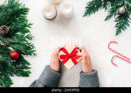 Kinderhände halten eine minimalistische Geschenkbox, die mit einem roten Band verziert ist. Tannenzweige, Kerzen und Zuckerrüben auf weißem strukturiertem Hintergrund. Bis Stockfoto