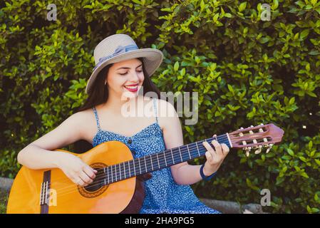 Junge Frau, die in der Sommersaison im Park Gitarre spielt und einen Hut verwendet Stockfoto