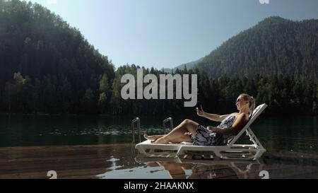 Frau, die mit einem Handheld-Film-Gimbal-Stabilisator für das Smartphone dreht. Mädchen ruhen, entspannen, liegen Sonnenbank auf Pier in Sonnenbrille, Selfie machen. Lady blogger bro Stockfoto