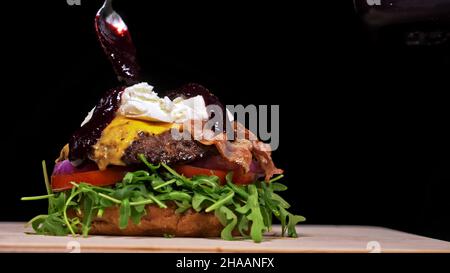 Handwerk Burger ist Kochen auf schwarzem Hintergrund. Bestehen: sauce, Rucola, Tomaten, roten Zwiebeln, Speck, rote Johannisbeeren Sauce, ricotta Cottage frischen Käse, Luft b Stockfoto