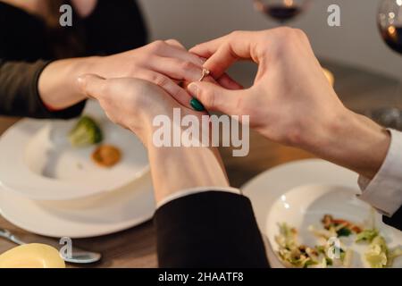 Ein Mann macht beim Abendessen einen Heiratsantrag an eine Frau, legt einen Verlobungsring auf den Finger eines Mädchens, ein Datum eines Paares in einem Restaurant, die Hände aus der Nähe Stockfoto