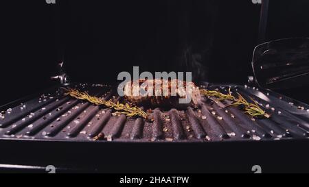 Köstliches, saftiges Steak-Kochen auf dem Grill. Braten Sie das Rindfleisch auf einem elektrischen Bräter, Rosmarin, schwarzem Pfeffer und Salz. Zeitlupe. Stockfoto