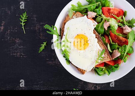 Frühstück - Toast mit Spiegeleiern und Tomaten-Salat mit Gemüse und Dorschleber auf dunklem Hintergrund. Gesundes Frühstück. Draufsicht Stockfoto