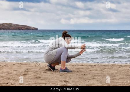 Frau, die ein Foto gemacht hat. Junge Fotografin, die sich am Sandstrand beugte und mit dem Smartphone eine Meeresaufnahme machte. Stockbild, Stockfoto